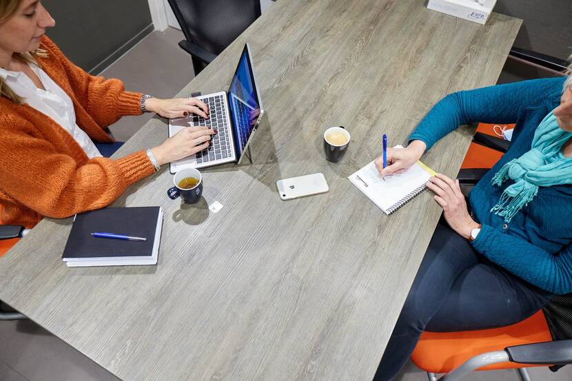 Twee vrouwen zitten aan tafel. Een van hen schrijft op een notitieblok, de ander typt op een laptop