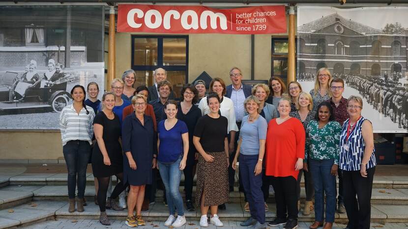 25 personen staan in een groep bij elkaar. Zij kijken naar en poseren voor de foto. Boven hen hangt een rode banner met de tekst Coram better changes for children since 1739