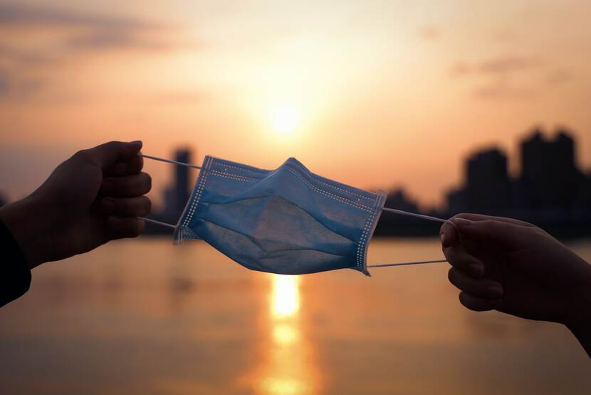 Twee handen hebben een mondkapje in hun hand. Op de achtergrond zie je een zonsondergang